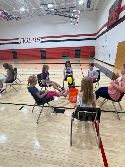 Students playing a teamwork game sitting in chairs and trying to kick a balloon into a bucket.