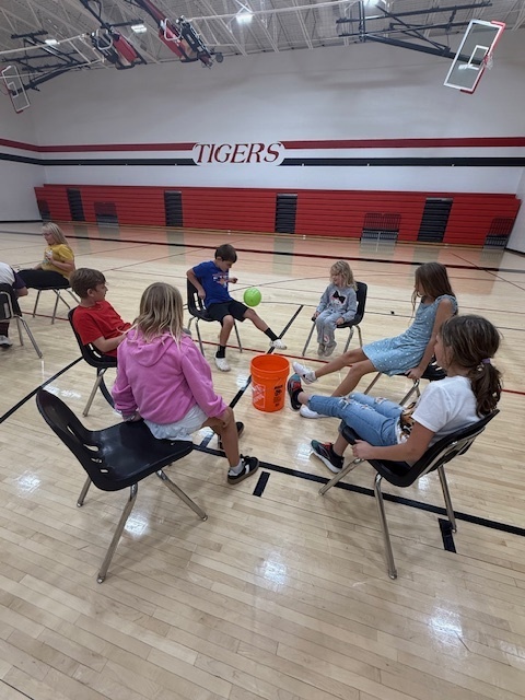 Students playing a teamwork game sitting in chairs and trying to kick a balloon into a bucket.