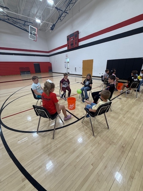 Students playing a teamwork game sitting in chairs and trying to kick a balloon into a bucket.