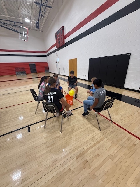 Students playing a teamwork game sitting in chairs and trying to kick a balloon into a bucket.