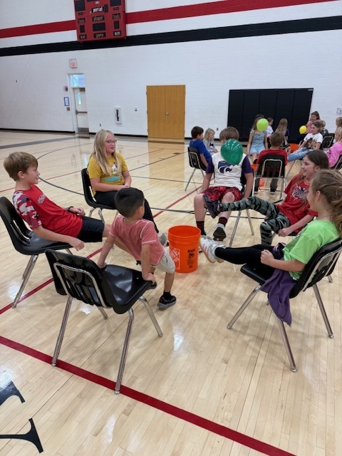 Students playing a teamwork game sitting in chairs and trying to kick a balloon into a bucket.
