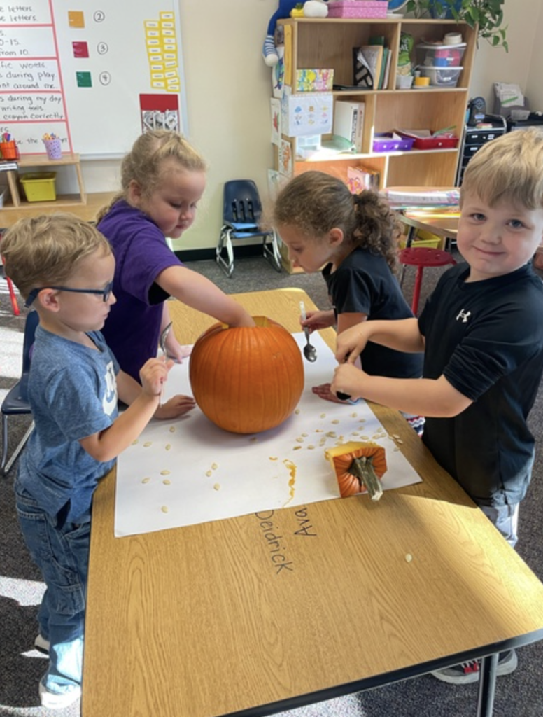 students at a table in a classroom smiling and discovering what the inside of a pumpkin looks and feels like