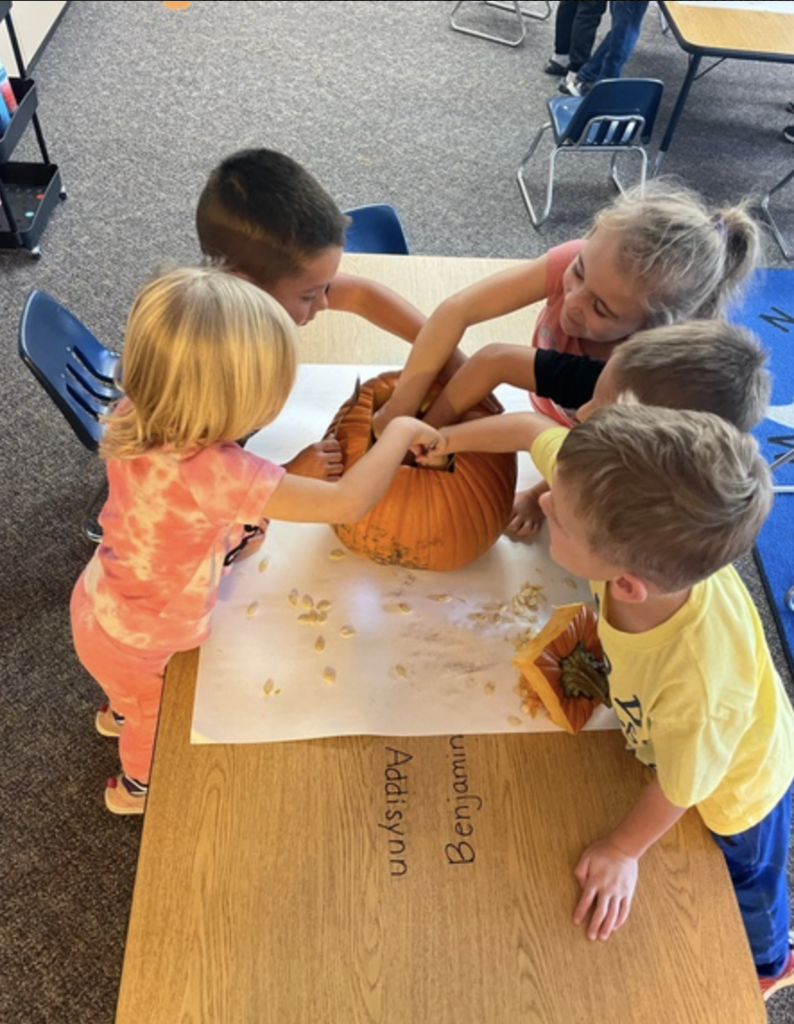 students at a table in a classroom smiling and discovering what the inside of a pumpkin looks and feels like