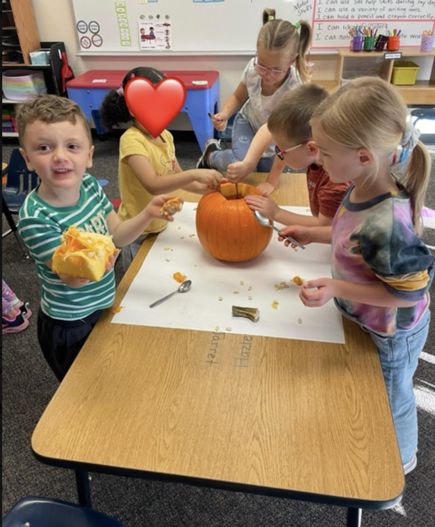 students at a table in a classroom smiling and discovering what the inside of a pumpkin looks and feels like