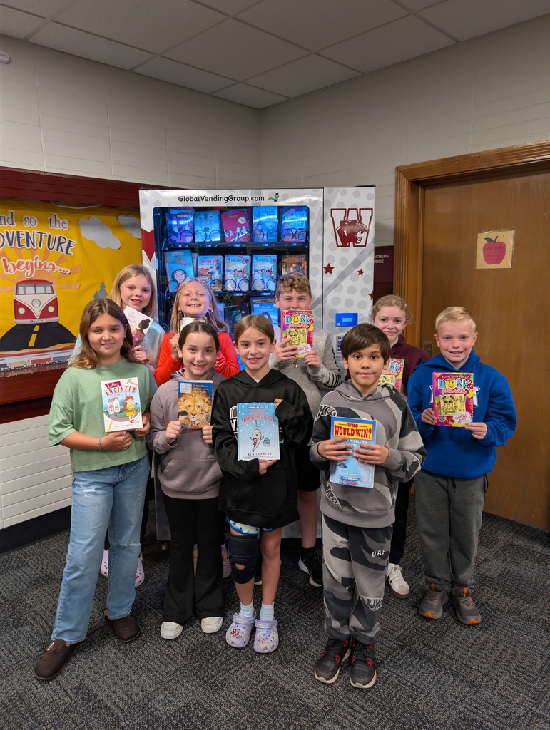 Several children in a room, holding books. One child holds up a book titled "The Tiger Who Came for Breakfast".