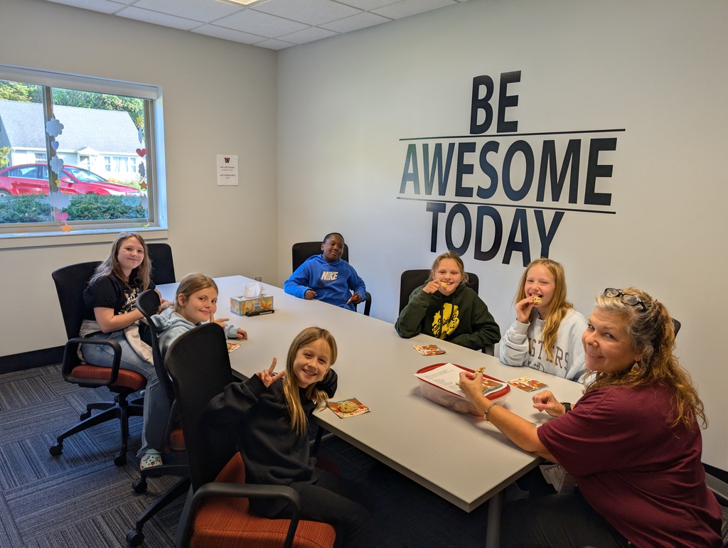 a group of people sit at a long table. each person has a cookie in their hand and is smiling.one student is showing a peace sign with her hand. there is a window in the background with a red car and a house. A wall reads, "Be Awesome Today."