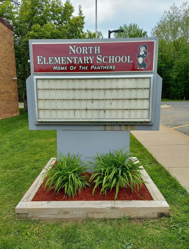 sign outside of building that says "north Elementary School home of the panthers" with 2 green plants below, blue skies with a cloud and trees in the back ground