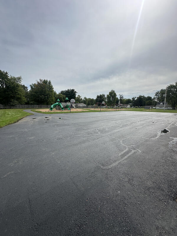 play ground with trees, green grass, basketball hoops, blacktop with puddles, sky with clouds a sun shining though. white houses in background and a telephone pole. everything is surrounded by green trees and a fence.