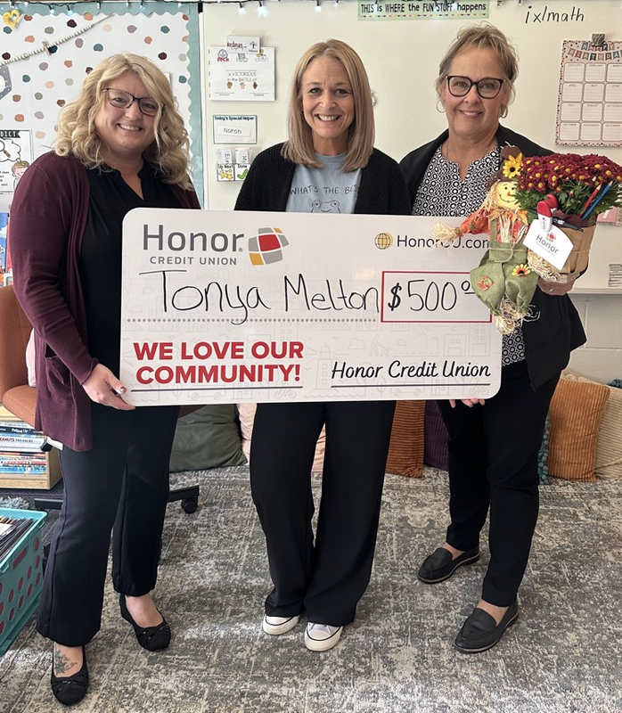Three adults pose in a classroom holding a $500 Honor Credit Union check made payable to Tonya Melton. One person holds a flower arrangement.