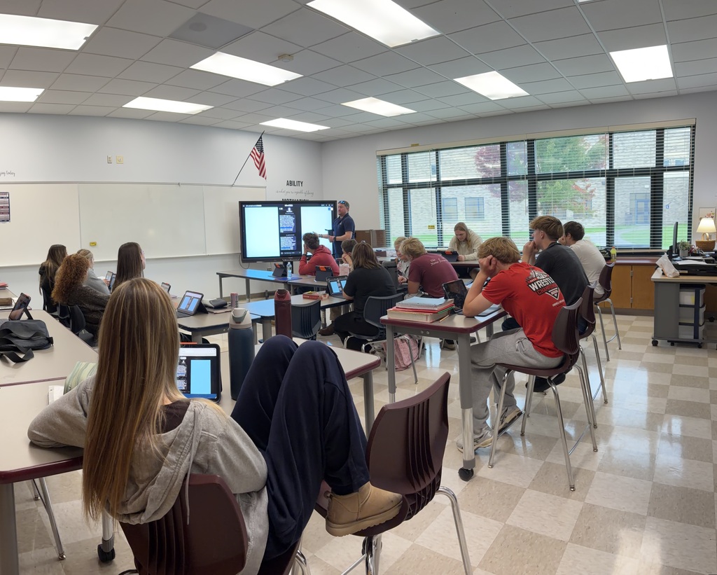 A classroom with students seated at desks, working on laptops. A teacher stands at a screen in the front.