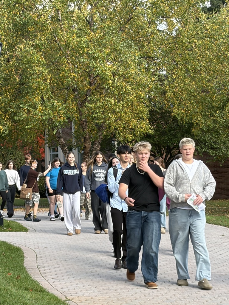 People walk on a brick path in a park. They are dressed casually. Trees with autumn leaves are in the background.