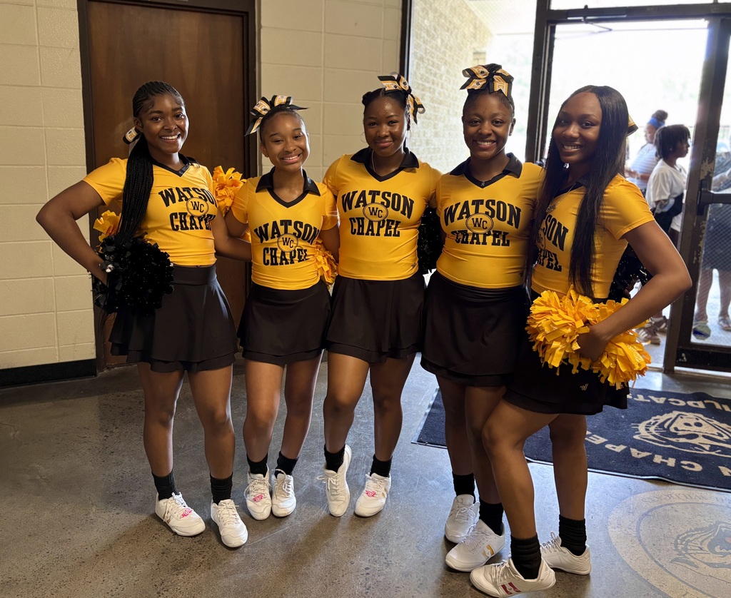 Five cheerleaders in matching yellow tops and black skirts, standing in a hallway with a door behind them.