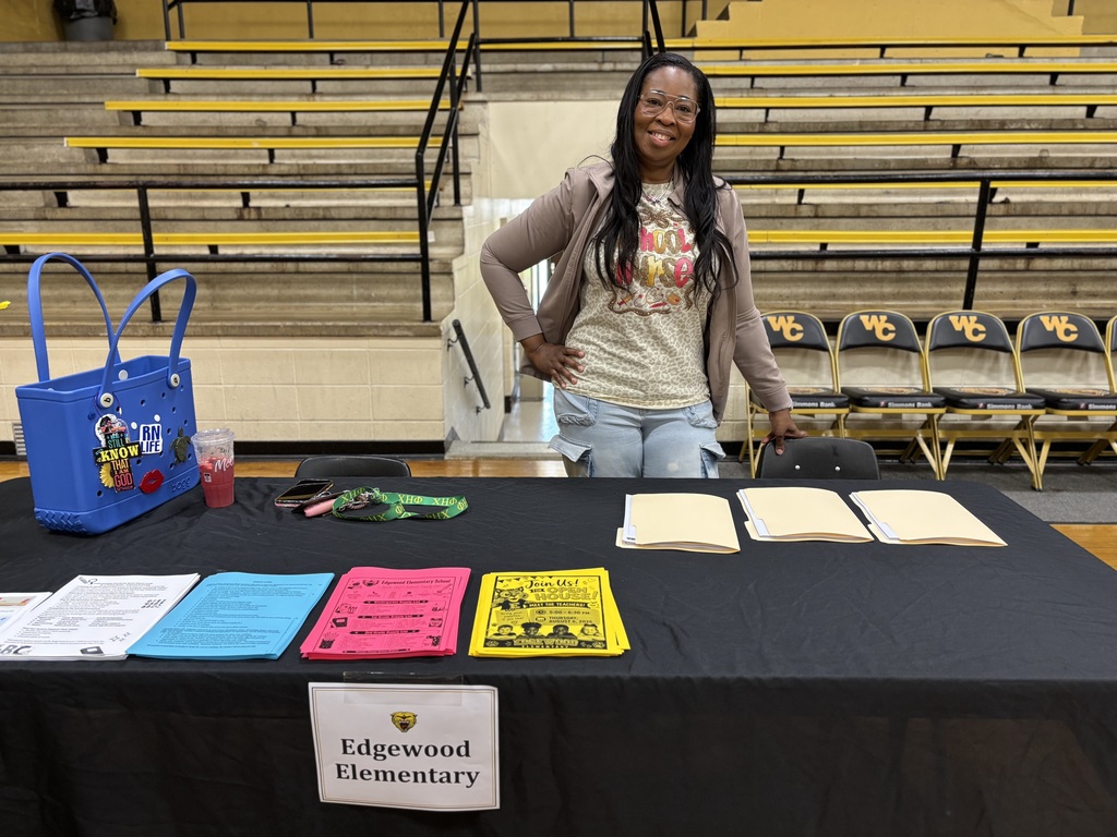Person at a table with various papers, a blue bag, and a sign reading "Edgewood Elementary" in a gym.