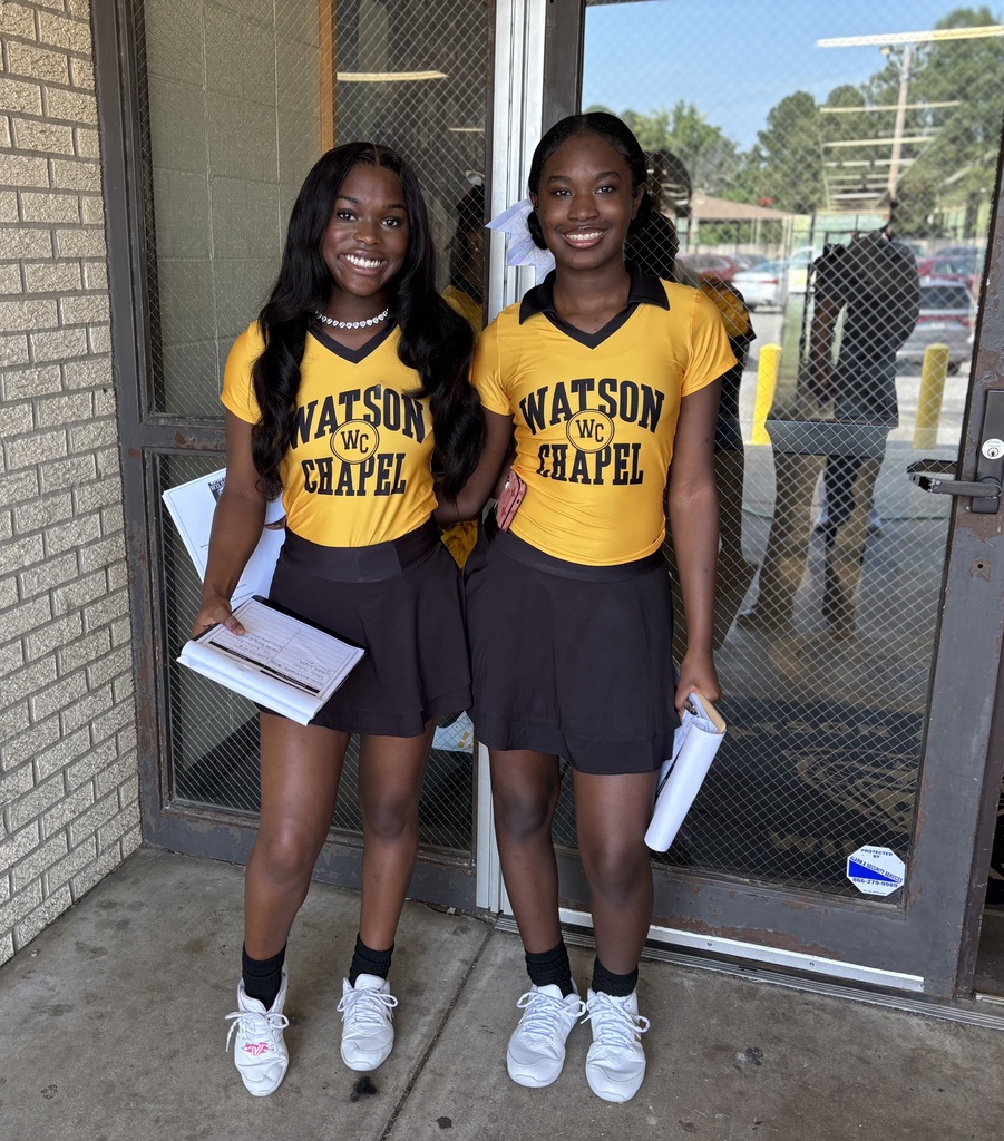 Two young women wearing yellow and black uniforms stand outside a building. They hold papers and wear sneakers.