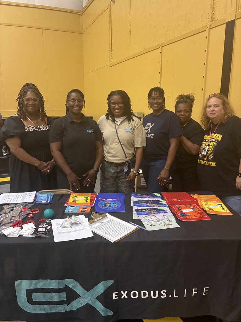 A group of adults stands behind a table covered with promotional materials and papers. The table reads "EXODUS.LIFE."
