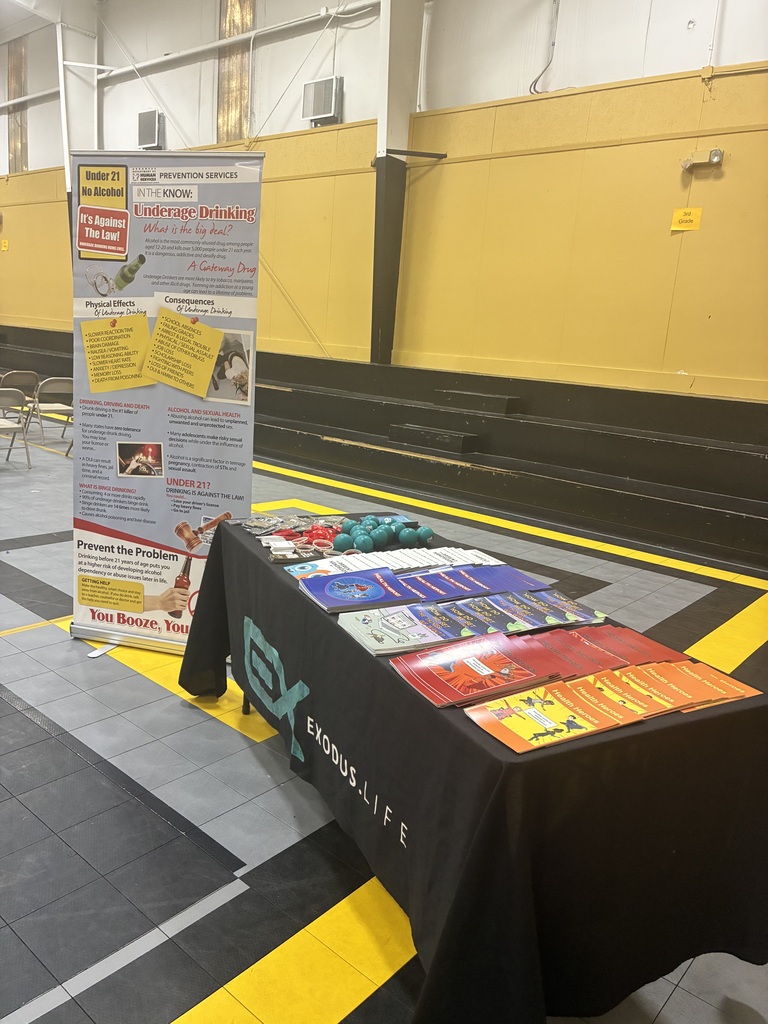 A table with a black tablecloth displays various materials, including pamphlets and books. A poster stands behind it, with yellow walls in the background.