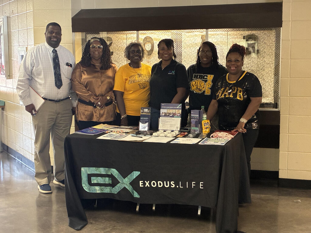Group of people standing behind a table with a black cover displaying "Exodus Life" text and logos, smiling.