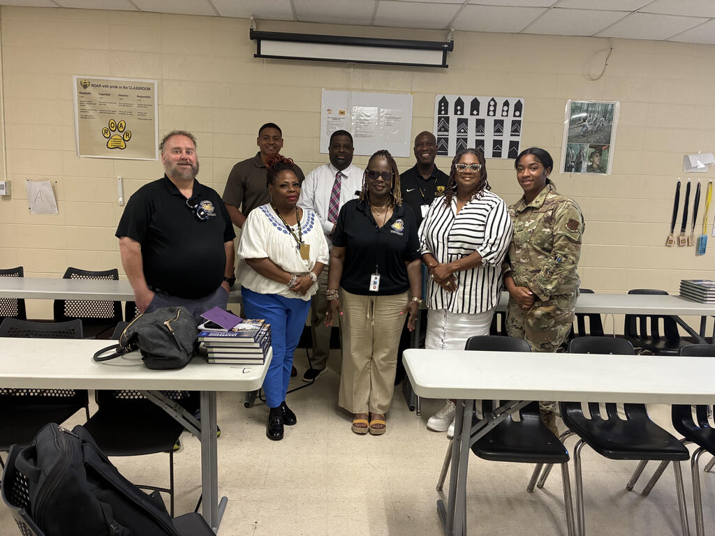 Group of adults in uniform and business attire standing together in a classroom, tables and chairs visible.