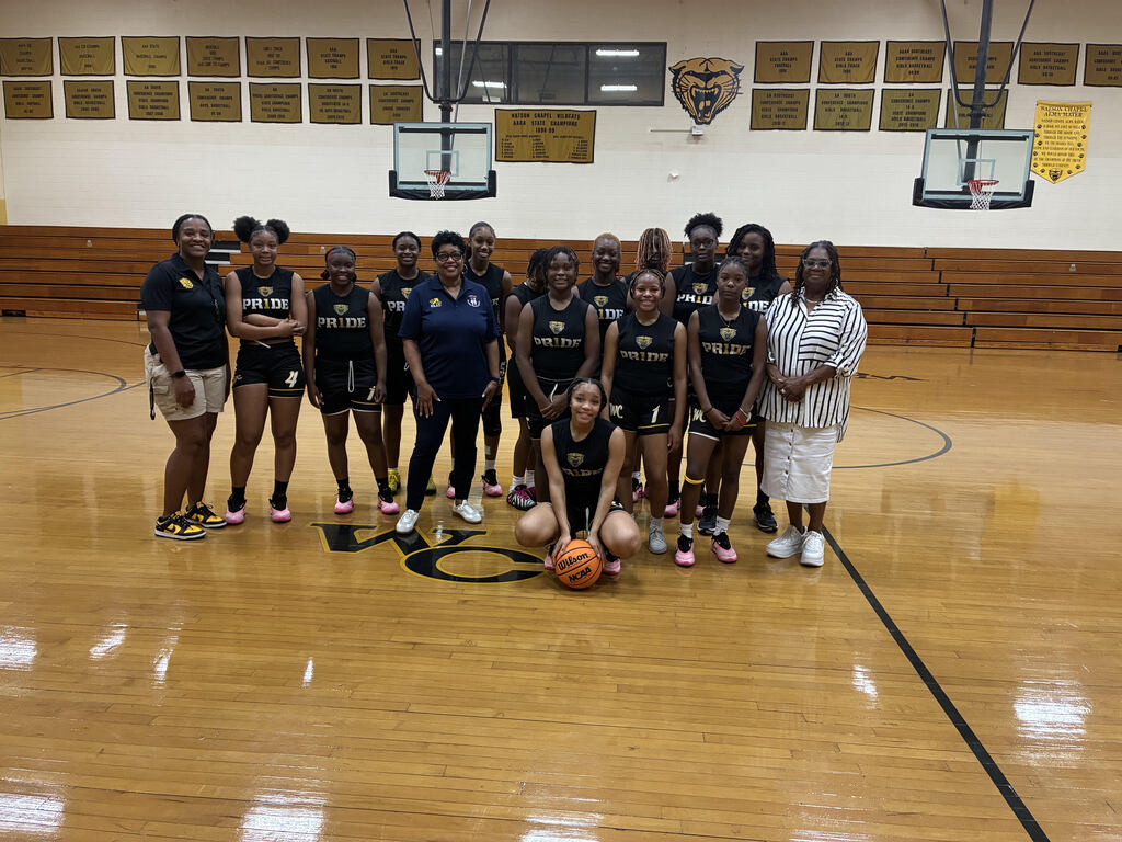 A team of women in black uniforms, sneakers, and a woman in white stand on a basketball court.