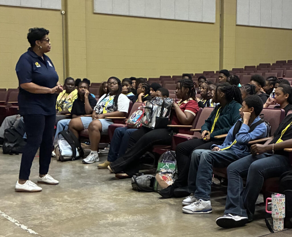 A woman stands at the front of a room, addressing a seated group. The audience is seated on chairs in rows.