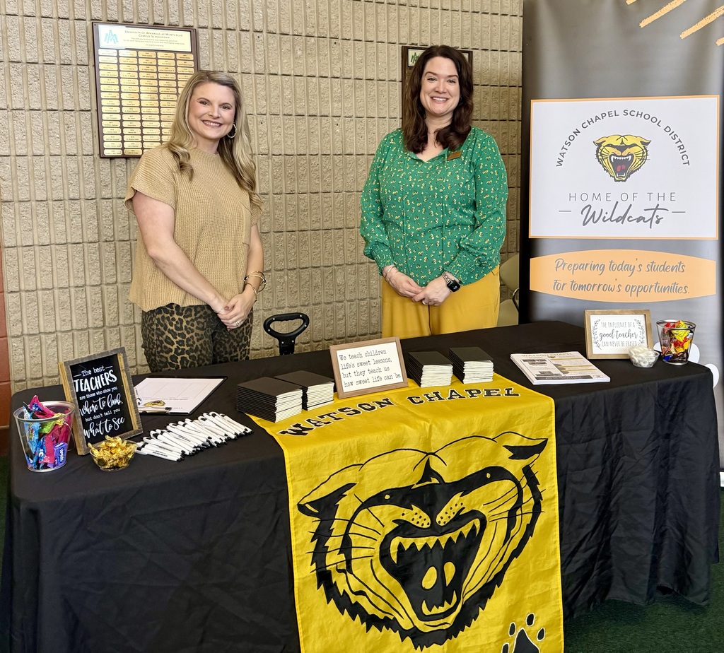 Two women stand behind a table with a yellow banner featuring a tiger. Papers, pencils, and other items are on the table. A sign reads "Home of the Wildcats".
