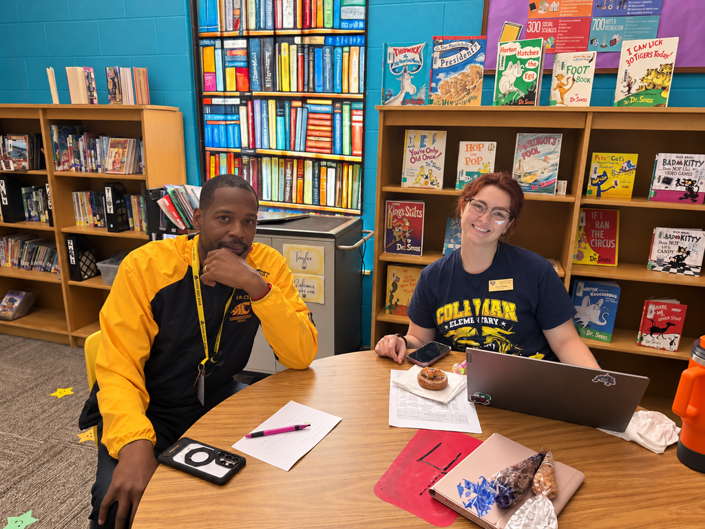Two adults seated at a table in a room with bookshelves, a laptop, pens, and a book.