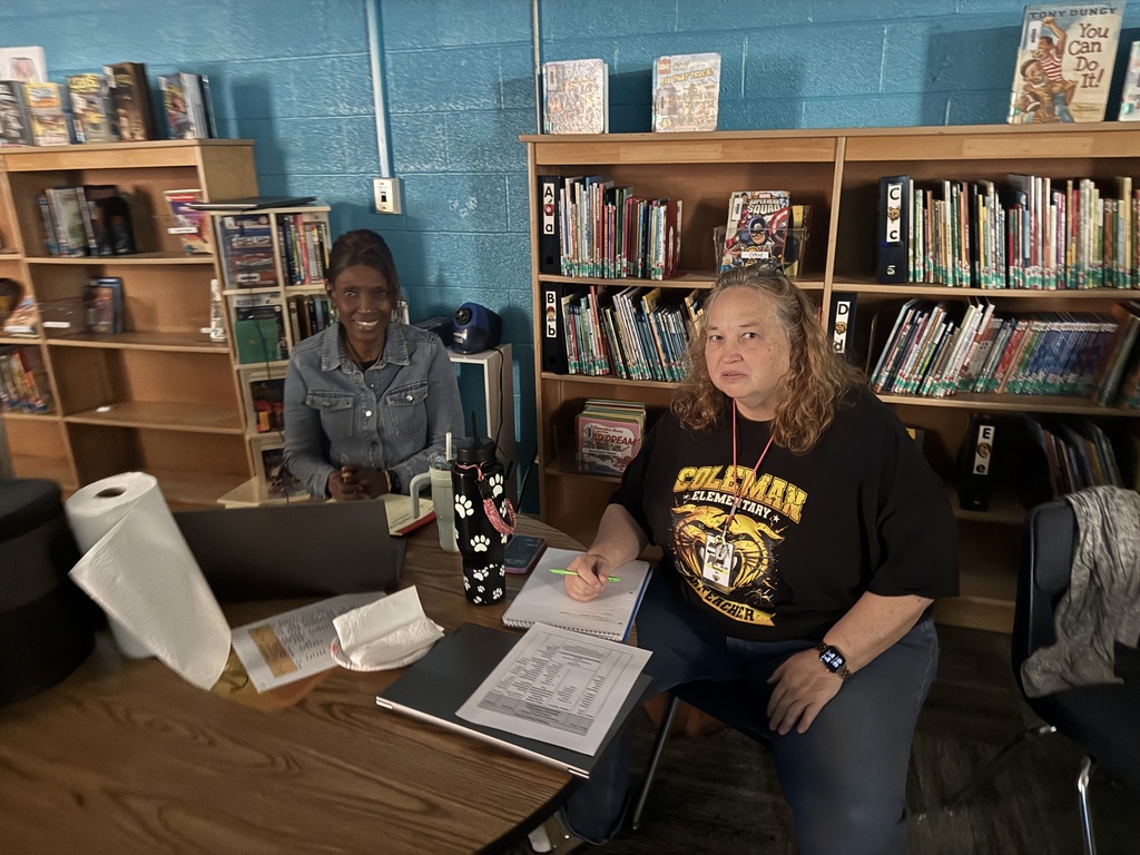 Two women sit at a desk, one writing on paper with a pen. They have a laptop, books, and a roll of paper in front of them. Behind them are two wooden bookcases filled with books.