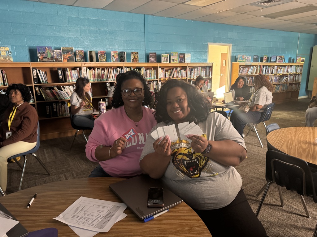 Two women sit at a table in a library, one holding cards, the other with a backpack.