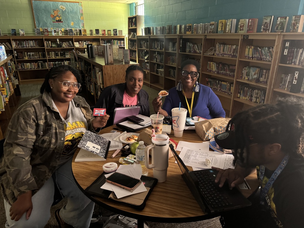 Four women sitting at a round table in a library, with food, books, and a laptop.