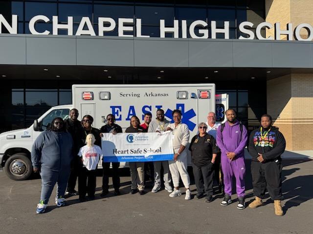 Group of people pose with banner in front of an ambulance at a school entrance.