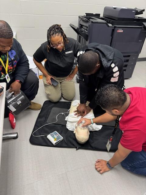 Four people gather around a dummy for training, two wearing IDs. One checks the dummy's mouth while another holds a tablet.