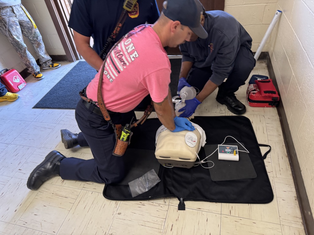 Two people kneeling on the floor, one wearing a pink shirt, practicing CPR on a dummy with an AED monitor nearby.