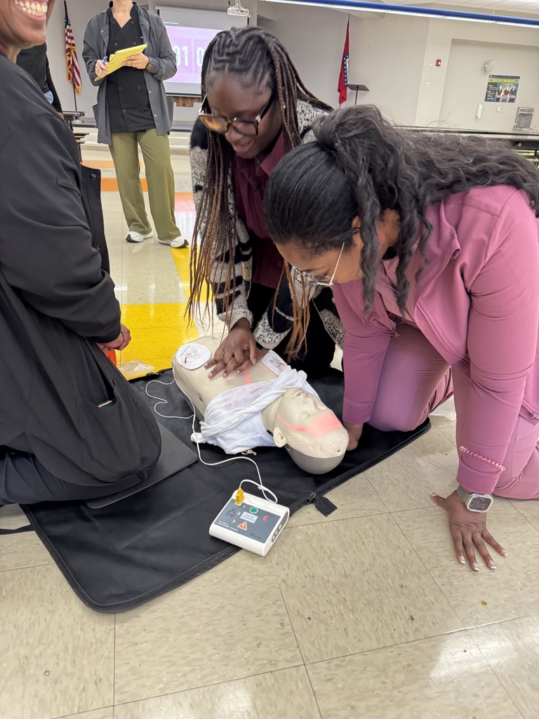 Three adults kneeling by a mannequin on the floor, one applying chest compressions. Another woman leans over the mannequin.