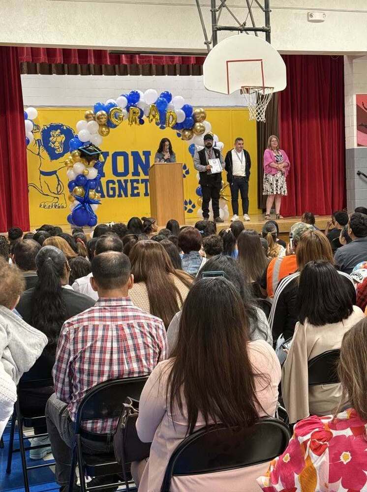 Graduation ceremony with audience members seated in the foreground and staff standing on a stage decorated with a balloon arch in the background