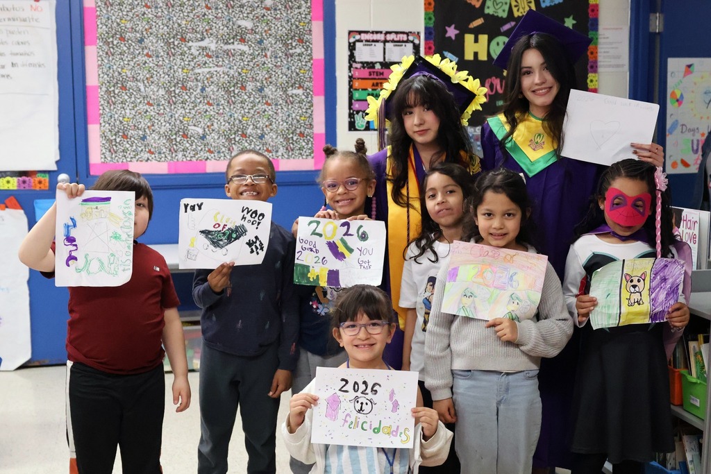 Elementary school students stand in a school hallway, proudly holding up colorful drawings they made as gifts for graduating high school seniors
