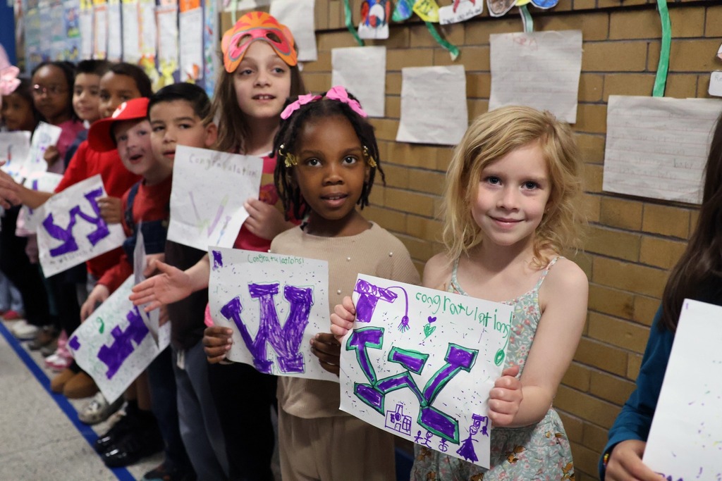 Elementary school students stand in a school hallway, proudly holding up colorful drawings they made as gifts for graduating high school seniors