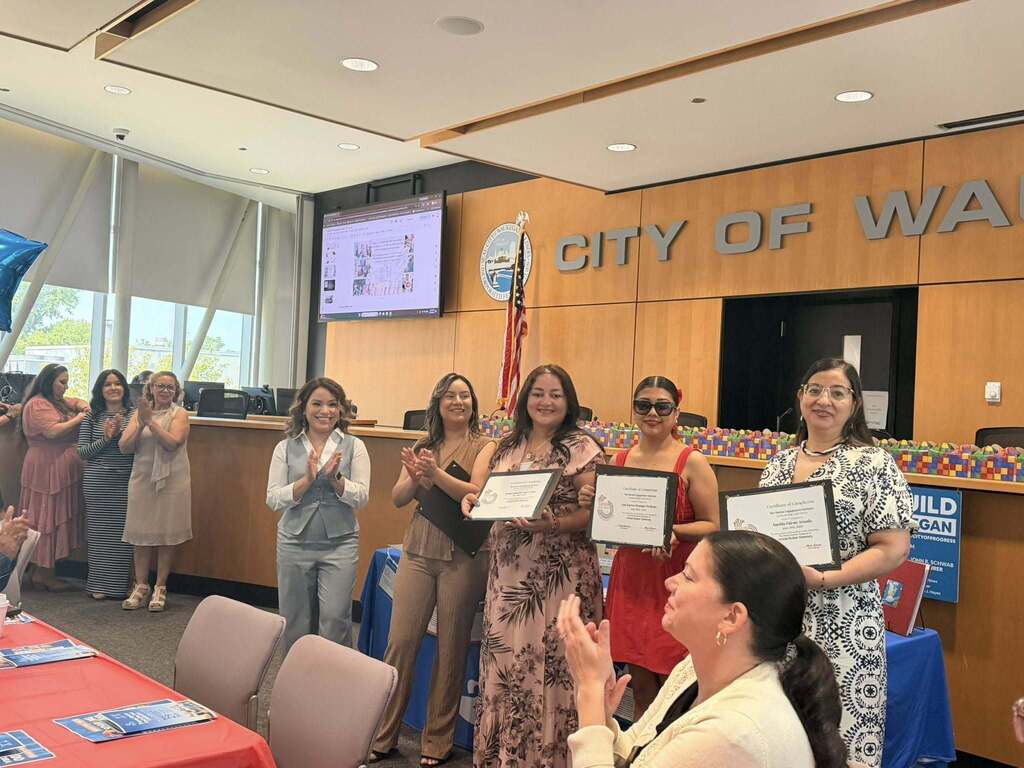 Three women stand together holding framed certificates while others applaud inside a city council chambers with 'CITY OF WAUKEGAN' visible on the wood-paneled wall