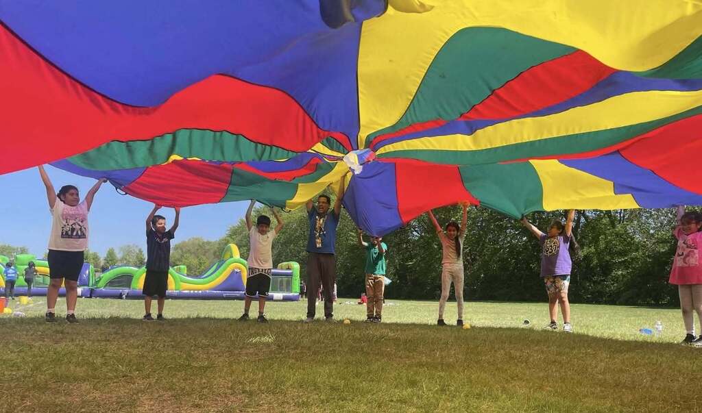group of children and an adult standing in a circle on a grassy field, lifting a large, vibrant, multi-colored play parachute high above their heads