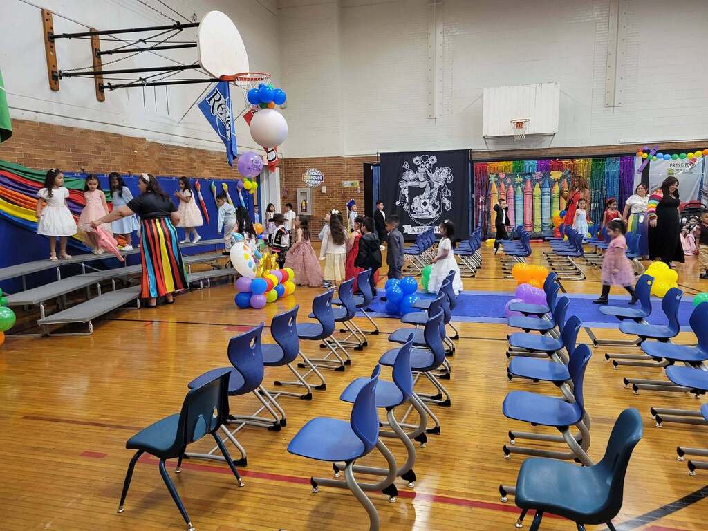 Pre-K students walking toward the stage during a graduation ceremony inside a gym filled with rows of chairs