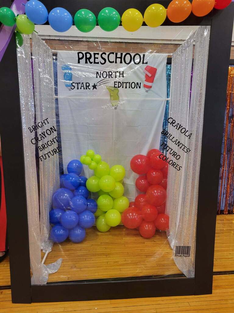 A colorful, crayon-box-themed photo booth for a preschool graduation featuring clusters of primary-colored balloons, silver curtains, a rainbow balloon arch, and a backdrop reading 'Preschool North Star Edition' in the file prek graduation