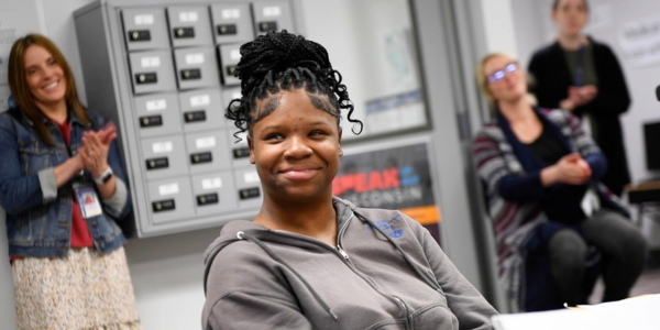 student smiling in foreground with staff clapping in background