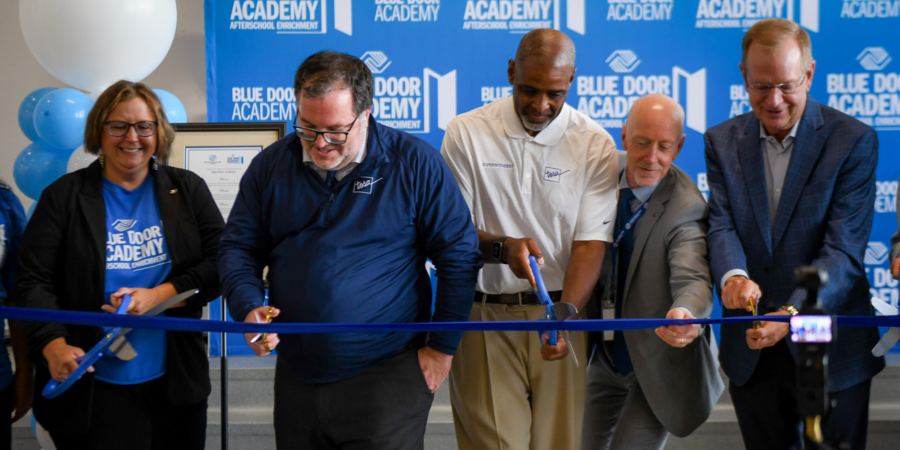 A group of school district and Boys & Girls Club leaders, including Ted Martin and Dr. Demond Means, cut a blue ribbon with giant blue scissors at a ribbon cutting ceremony for Blue Door Academy at Underwood Elementary School.