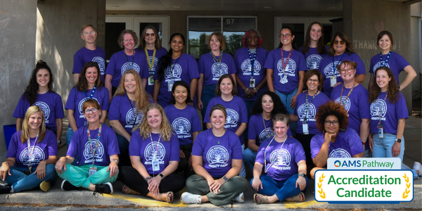 The Wauwatosa Montessori School staff poses in front of the school building, wearing matching purple t-shirts and smiling at the camera.