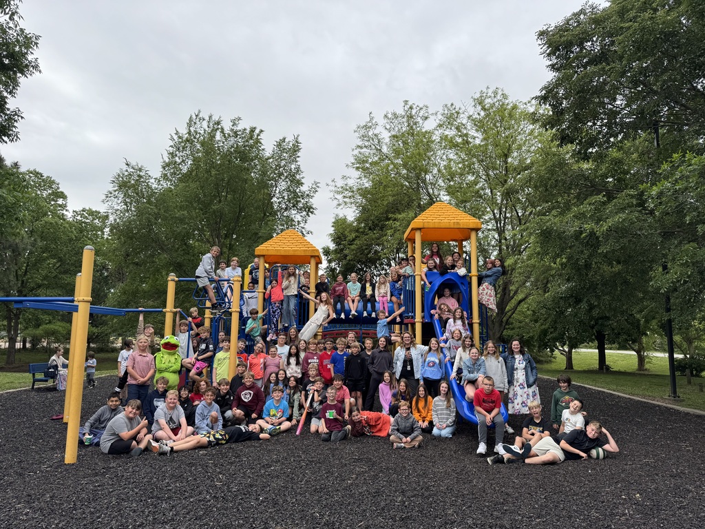 5th grade students, some seated and some standing, pose for a photo in a park. Playground equipment is in the background.