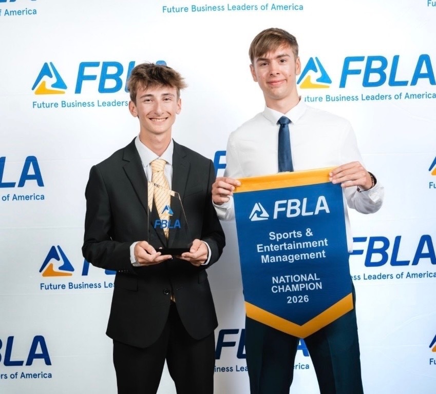 Two men hold a blue banner with "FBLa National Champion 2026" in front of a banner with the FBLa logo.