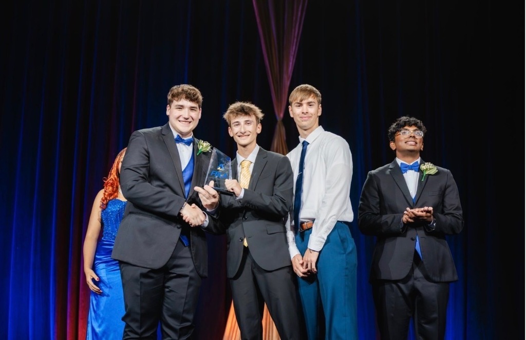 Five men in suits and ties stand on a stage; one holds an award, with a person in blue behind them and a blue curtain in the background.