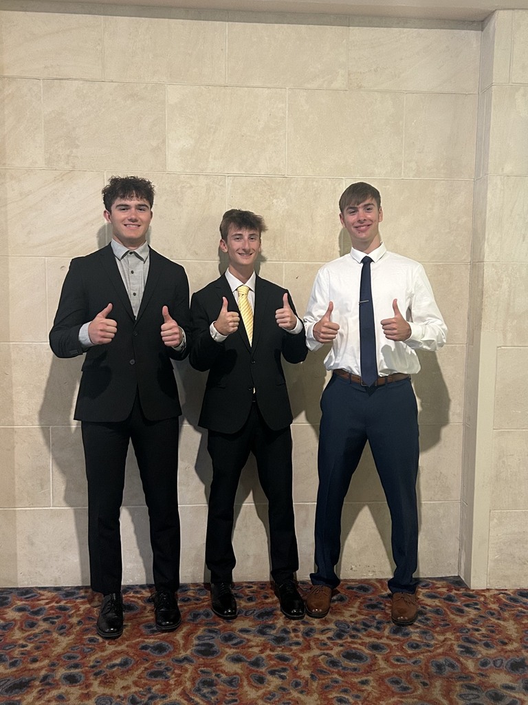 Three young men in formal attire stand next to each other, giving thumbs up. They are in a room with a patterned floor and a beige wall.