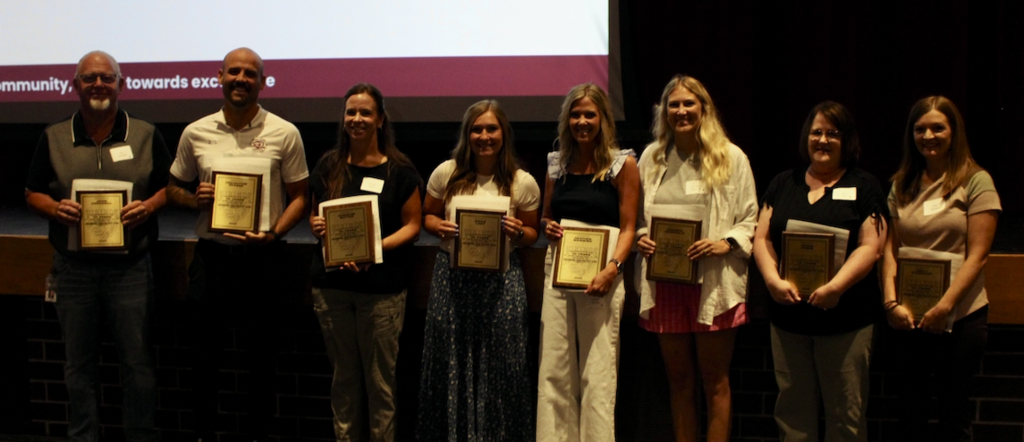 A group of eight women and two men pose with awards in hand in front of a projector screen.
