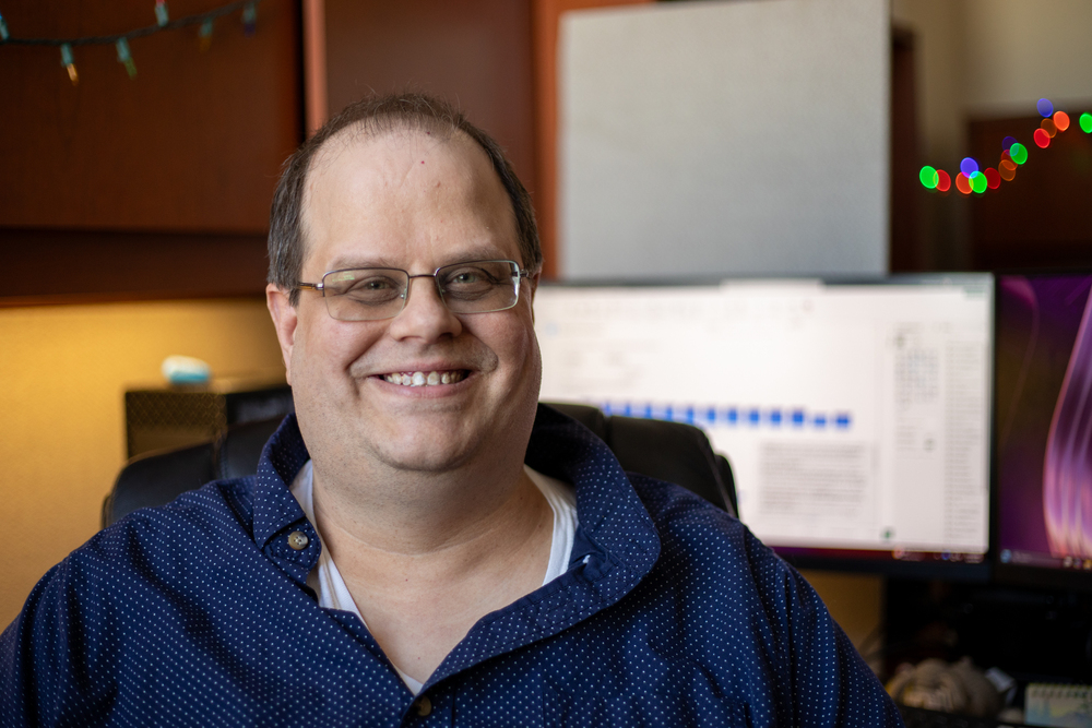 Luke Newhart sits in front of his computer monitor and smiles.