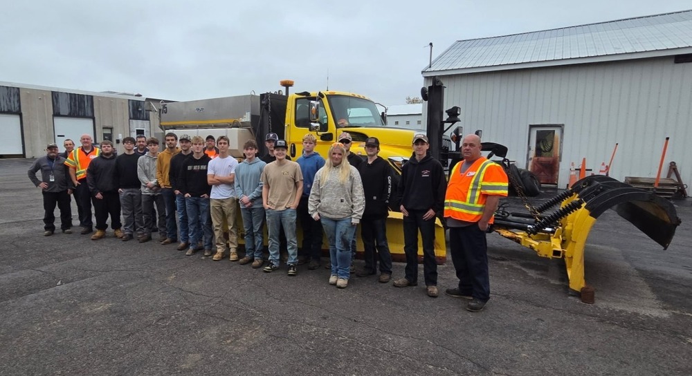 About 15 WTCC students stand in a row in front of a yellow snowplow truck outside in a gravel parking lot.